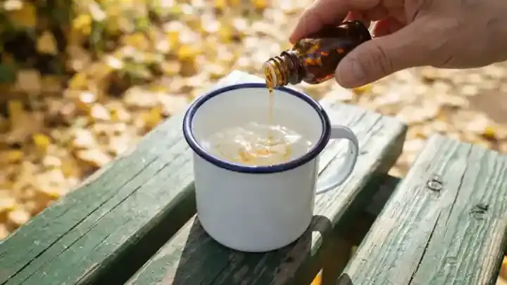 A single drop of amber herbal extract falling into a white enamel camp cup of water on a park bench, faint color swirling, slow zoom